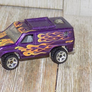 May include: A purple toy van with orange and yellow flame decals. The van has black wheels with silver rims and a roof rack. The Hot Wheels logo is visible on the side of the van. The toy is on a wooden surface.
