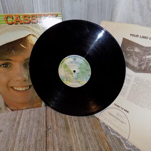 May include: A black vinyl record with a colorful label is centered in the image. To the left is a record album cover featuring a smiling man wearing a white hat. A paper insert with the text "YOUR LIMO IS READY..." is visible.