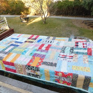 May include: A patchwork quilt made from a variety of colorful fabrics, including red, blue, yellow, green, and white. The quilt is laid out on a wooden picnic table.