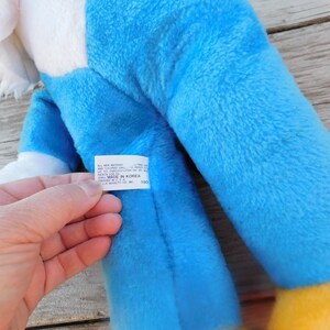 May include: Blue and white plush toy with a yellow beak and feet. The toy has a tag that reads "All New Material, 100% Colored Silk, No. 40,  Made in Korea,  Packed in U.S.A.,  Finished in U.S.A.,  J.L.  Marchetti, Inc., 1100".