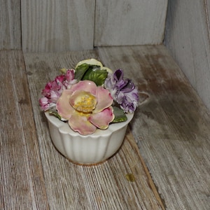 May include: A white porcelain flower pot with a ruffled rim, decorated with a bouquet of pink, purple, and yellow flowers.