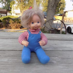 Puede incluir: Una mu&ntilde;eca vintage con cabello rubio, vistiendo una camisa a cuadros rojos y blancos y un mono azul. La mu&ntilde;eca est&aacute; sentada sobre una superficie de madera.