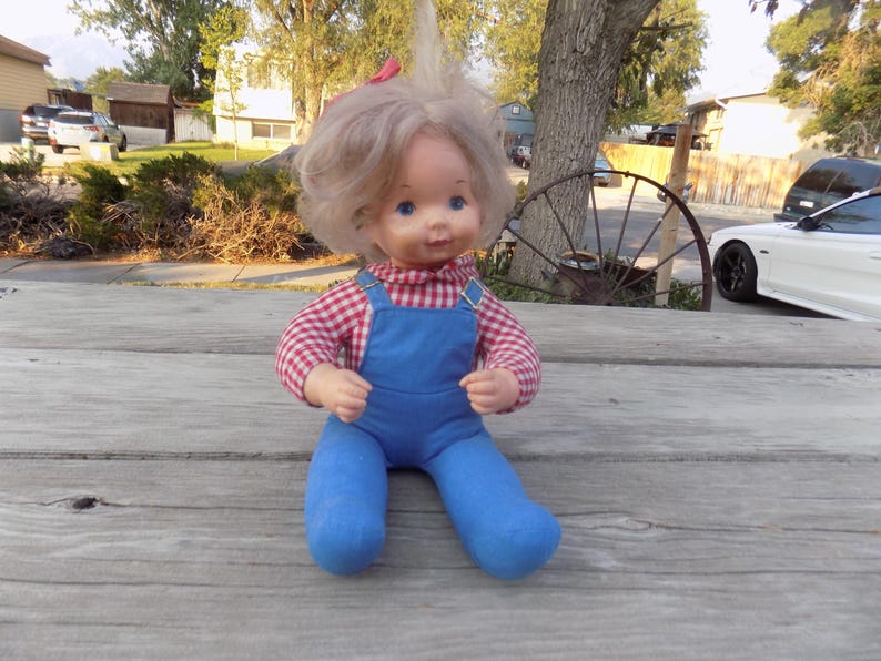 Puede incluir: Una mu&ntilde;eca vintage con cabello rubio, vistiendo una camisa a cuadros rojos y blancos y un mono azul. La mu&ntilde;eca est&aacute; sentada sobre una superficie de madera.