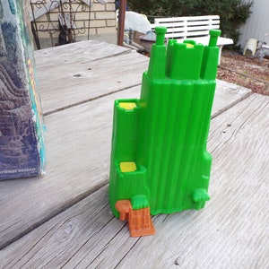 May include: A green plastic toy castle with yellow details and an orange base. The castle has a tall tower and multiple smaller towers. The castle is sitting on a wooden surface.