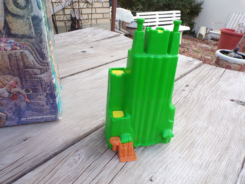 May include: A green plastic toy castle with yellow details and an orange base. The castle has a tall tower and multiple smaller towers. The castle is sitting on a wooden surface.