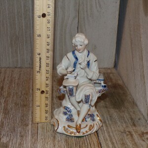 Pu&ograve; includere: Una statuetta in porcellana di un uomo seduto su una panchina, che tiene un uccello in mano. La statuetta &egrave; dipinta in blu e bianco con accenti dorati.