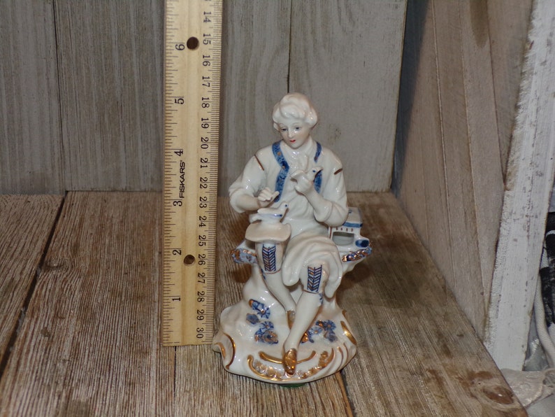 Pu&ograve; includere: Una statuetta in porcellana di un uomo seduto su una panchina, che tiene un uccello in mano. La statuetta &egrave; dipinta in blu e bianco con accenti dorati.