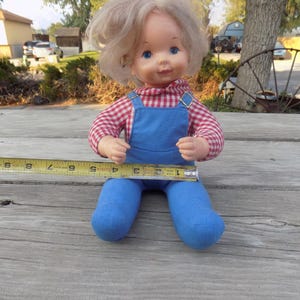 Puede incluir: Una mu&ntilde;eca vintage con cabello rubio y ojos azules que lleva una camisa a cuadros rojos y blancos y un mono azul. La mu&ntilde;eca est&aacute; sentada sobre una superficie de madera.