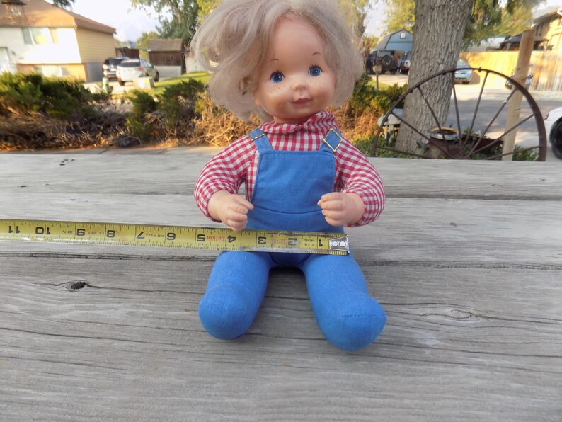 Puede incluir: Una mu&ntilde;eca vintage con cabello rubio y ojos azules que lleva una camisa a cuadros rojos y blancos y un mono azul. La mu&ntilde;eca est&aacute; sentada sobre una superficie de madera.