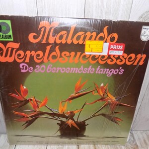 May include: A brown vinyl record album cover with the title "Malando Wereldsuccessen" in orange and yellow lettering. The album cover features a floral arrangement of orange and yellow bird of paradise flowers in a brown pot. The text "De 30 beroemdste tango's" is printed below the title. The album cover also features a sticker with the text "Prijs 21.95" in red and white lettering.