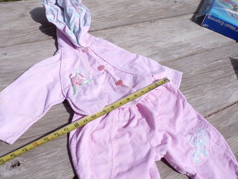 Puede incluir: Un conjunto de beb&eacute; de pana rosa con una chaqueta con capucha y pantalones. La chaqueta tiene un dise&ntilde;o floral y botones. Los pantalones tienen un dise&ntilde;o floral en una pierna. Una cinta m&eacute;trica se coloca sobre el conjunto.