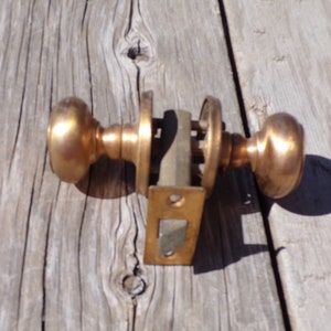 May include: A vintage brass door knob set with two round knobs and a square latch plate. The knobs are a shiny brass color and the latch plate is a slightly duller brass color.