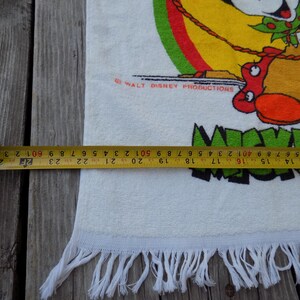 Peut inclure: Serviette en coton blanche avec un dessin color&eacute; de Mickey Mouse en tenue de cow-boy. La serviette, d'environ 63 cm de large, a une bordure &agrave; franges et le texte "Mickey Mouse" en vert. Le logo Walt Disney Productions est &eacute;galement visible.