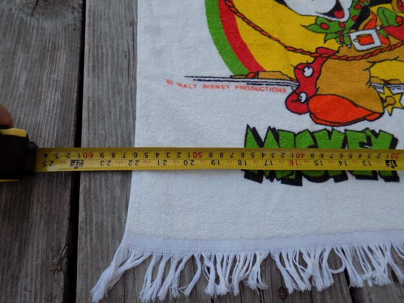 Peut inclure: Serviette en coton blanche avec un dessin color&eacute; de Mickey Mouse en tenue de cow-boy. La serviette, d'environ 63 cm de large, a une bordure &agrave; franges et le texte "Mickey Mouse" en vert. Le logo Walt Disney Productions est &eacute;galement visible.