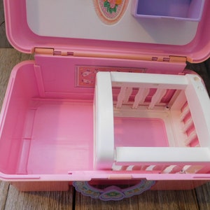 Puede incluir: Un juego de pl&aacute;stico rosa en forma de maleta con una cuna blanca en el interior. La maleta tiene un dise&ntilde;o floral en la parte delantera y un peque&ntilde;o estante morado en la parte superior.