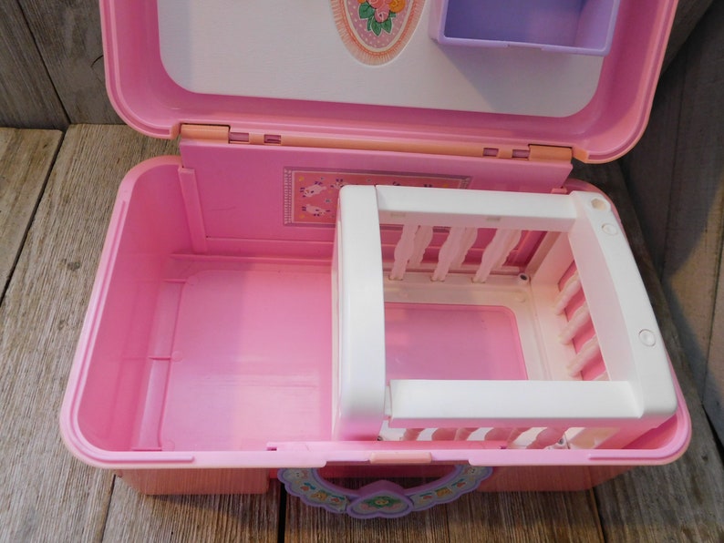Puede incluir: Un juego de pl&aacute;stico rosa en forma de maleta con una cuna blanca en el interior. La maleta tiene un dise&ntilde;o floral en la parte delantera y un peque&ntilde;o estante morado en la parte superior.