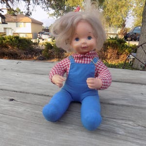 Puede incluir: Una mu&ntilde;eca vintage con cabello rubio que lleva una camisa a cuadros rojos y blancos y un mono azul. La mu&ntilde;eca est&aacute; sentada sobre una superficie de madera.