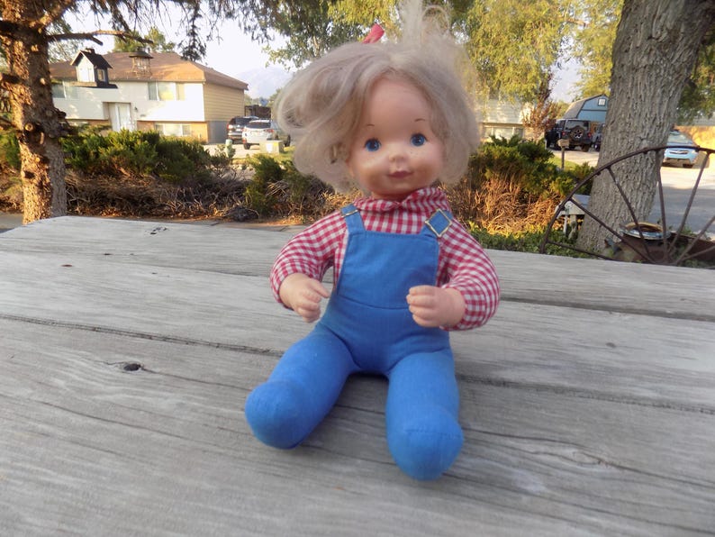 Puede incluir: Una mu&ntilde;eca vintage con cabello rubio que lleva una camisa a cuadros rojos y blancos y un mono azul. La mu&ntilde;eca est&aacute; sentada sobre una superficie de madera.