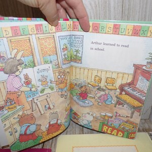 May include: An open children's book with colorful illustrations of animals and a classroom setting. The book features the text "Why are books delicious things?" and "Jump!" The book also has a colorful alphabet border.