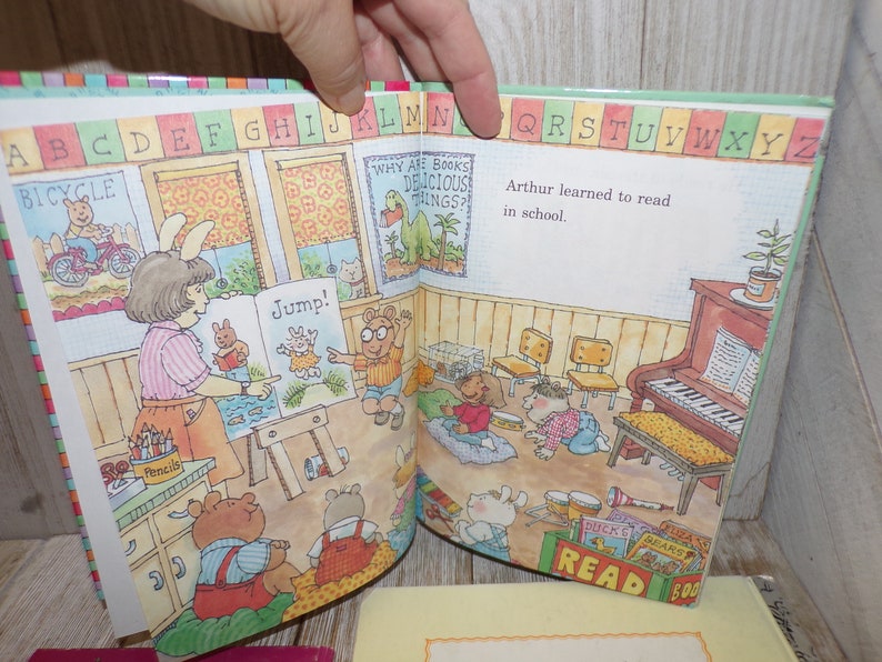 May include: An open children's book with colorful illustrations of animals and a classroom setting. The book features the text "Why are books delicious things?" and "Jump!" The book also has a colorful alphabet border.