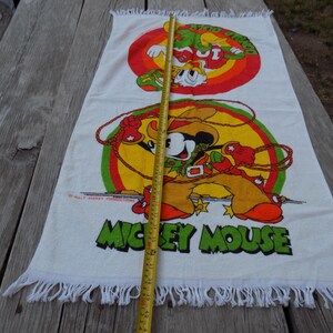 Peut inclure: Serviette de plage rectangulaire blanche avec un dessin anim&eacute; color&eacute; de Mickey Mouse en cow-boy avec un lasso et un second dessin anim&eacute; de Dingo. La serviette a un bord &agrave; franges et les mots "MICKEY MOUSE" en vert.