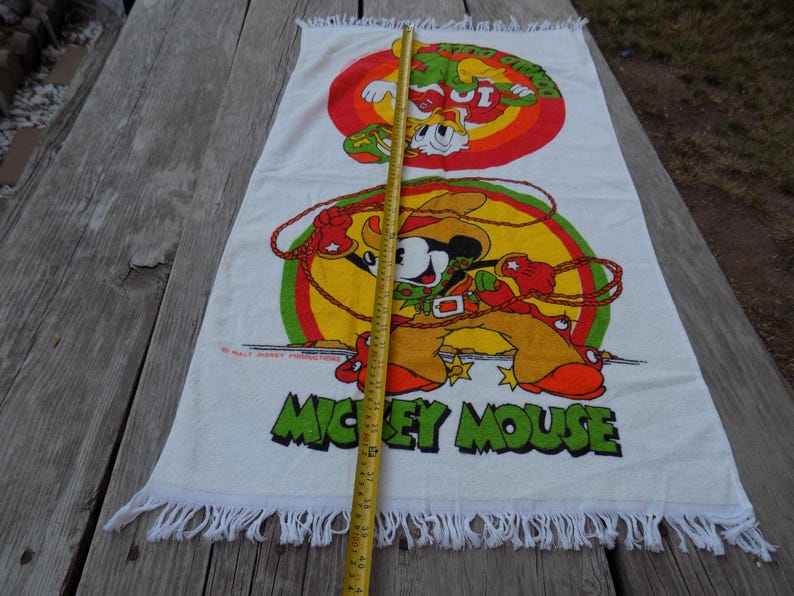 Peut inclure: Serviette de plage rectangulaire blanche avec un dessin anim&eacute; color&eacute; de Mickey Mouse en cow-boy avec un lasso et un second dessin anim&eacute; de Dingo. La serviette a un bord &agrave; franges et les mots "MICKEY MOUSE" en vert.