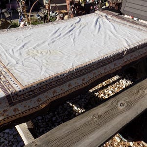 May include: A rectangular tablecloth with a cream-colored center and a decorative border. The border features a repeating pattern of floral and geometric elements in brown, tan, and blue. The tablecloth is draped over a wooden surface.