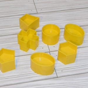 May include: A collection of bright yellow geometric shape cookie cutters. The shapes include a square, a cross, a circle, an oval, a pentagon, a hexagon, and a rounded rectangle. The cutters are made of plastic and are arranged on a light wood surface.