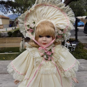 May include: A porcelain doll with blonde hair and blue eyes, wearing a cream-colored dress and bonnet. The dress features pleated fabric, lace trim, and pink ribbon accents. The bonnet is adorned with lace and small pink roses.