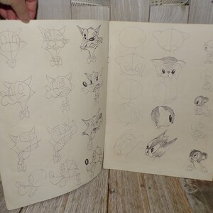 May include: Open book with pencil sketches of cartoon animal faces. The drawings show the steps of creating the cartoon faces, with each step outlined in pencil. The animals include cats, dogs, and other cartoon characters.