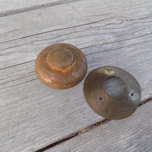 May include: Two antique bronze hardware pieces. One is a round knob with a smaller top, and the other is a circular plate with a hole in the center. Both pieces have a weathered, aged appearance.