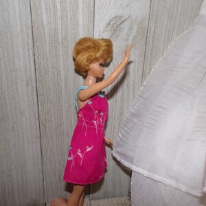 May include: A vintage Barbie doll wearing a pink dress with a floral pattern and a light blue trim. The doll has blonde hair styled in a bouffant and blue eyes. The doll is standing on a wooden surface.