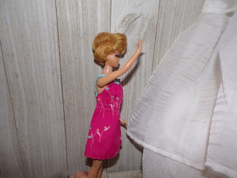 May include: A vintage Barbie doll wearing a pink dress with a floral pattern and a light blue trim. The doll has blonde hair styled in a bouffant and blue eyes. The doll is standing on a wooden surface.