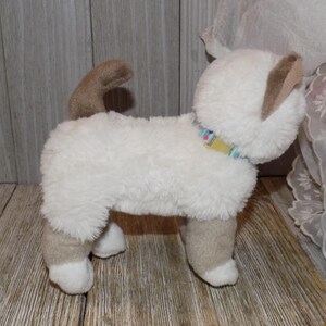 May include: A plush cat toy in a standing position. The toy is primarily white with tan accents on the ears, legs, and tail. It has a colorful collar and is made of soft, fuzzy material. The toy is designed to resemble a Siamese cat.