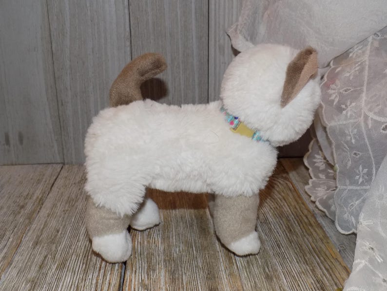 May include: A plush cat toy in a standing position. The toy is primarily white with tan accents on the ears, legs, and tail. It has a colorful collar and is made of soft, fuzzy material. The toy is designed to resemble a Siamese cat.