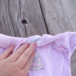 May include: A pink baby onesie with a white bunny rabbit wearing a pink dress and holding a teacup. The onesie has a white tag that says "Carter's" on the front.