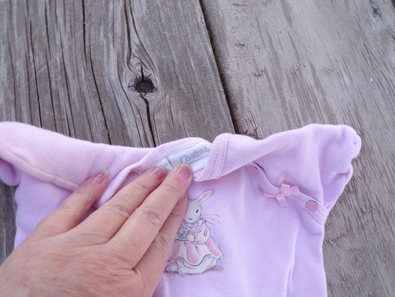 May include: A pink baby onesie with a white bunny rabbit wearing a pink dress and holding a teacup. The onesie has a white tag that says "Carter's" on the front.