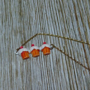 May include: A gold chain necklace with three cupcake charms. The cupcakes are white with orange frosting and pink cherries on top.