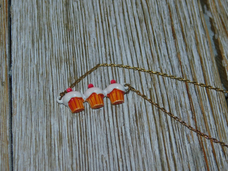 May include: A gold chain necklace with three cupcake charms. The cupcakes are white with orange frosting and pink cherries on top.