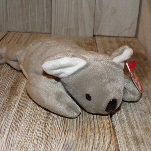 May include: A gray and white plush koala bear toy with a red and white beanie baby tag. The koala is lying on its belly with its head turned to the side.