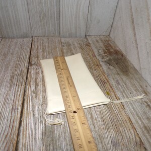 Puede incluir: Una bolsa de tela blanca con cierre de cord&oacute;n, medida con una regla de madera. La bolsa mide aproximadamente 15 cm de largo.
