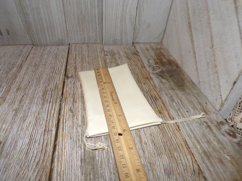 Puede incluir: Una bolsa de tela blanca con cierre de cord&oacute;n, medida con una regla de madera. La bolsa mide aproximadamente 15 cm de largo.