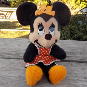 May include: A plush Minnie Mouse doll with black fur, large black ears, and an orange bow. She wears a red and white polka dot dress and orange fluffy shoes. The doll is sitting on a wooden surface.