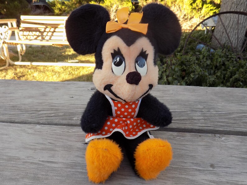 May include: A plush Minnie Mouse doll with black fur, large black ears, and an orange bow. She wears a red and white polka dot dress and orange fluffy shoes. The doll is sitting on a wooden surface.
