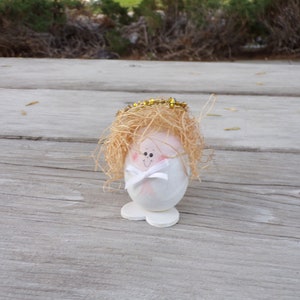 May include: A decorative angel figurine made from an eggshell. The angel has a painted face, straw hair, a gold halo, and a white bow. The figurine is standing on a white base. The background is a wooden surface.