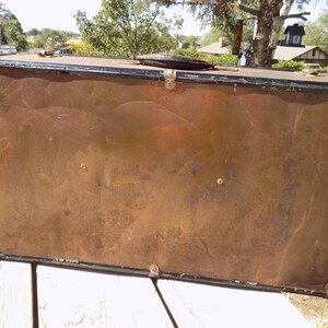 May include: A vintage trunk with a copper-colored exterior and black trim. The trunk has metal corner protectors and a leather handle. The trunk is rectangular and appears to be made of metal.