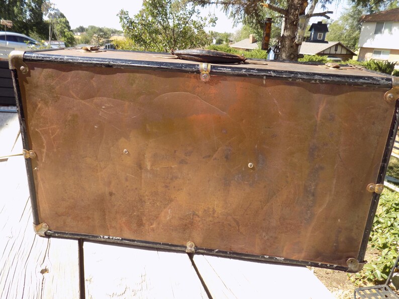 May include: A vintage trunk with a copper-colored exterior and black trim. The trunk has metal corner protectors and a leather handle. The trunk is rectangular and appears to be made of metal.