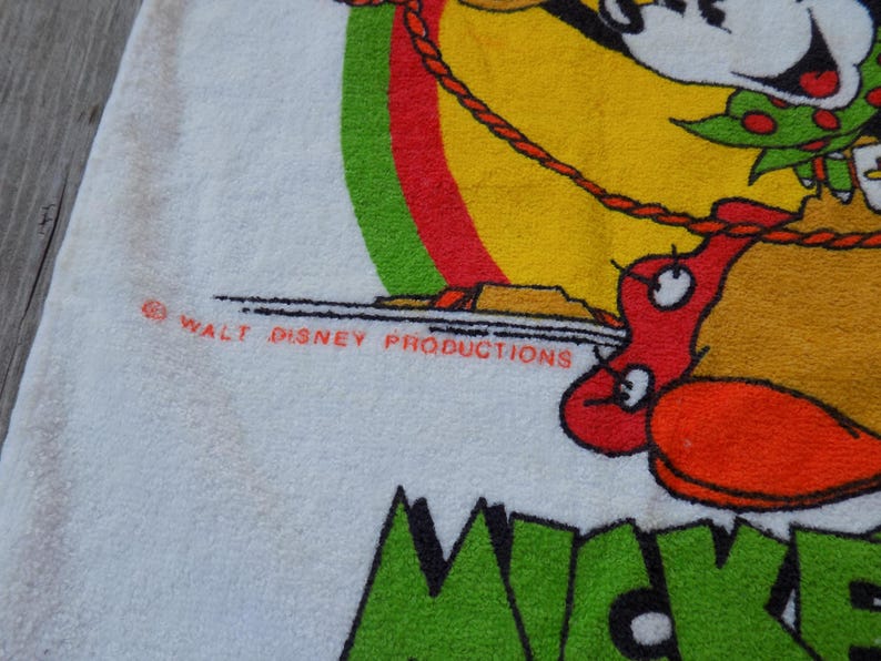 Peut inclure: Serviette blanche avec un motif Mickey Mouse color&eacute;. Le motif comprend Mickey Mouse, un fond rouge et jaune, et le texte "Mickey". La serviette porte &eacute;galement le texte "WALT DISNEY PRODUCTIONS".