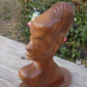 May include: A carved wooden bust of a person with closed eyes and a serene expression. The sculpture features detailed hair and a smooth, polished finish. The bust is a warm brown color and rests on a circular base.