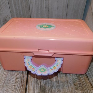 Puede incluir: Una caja de almacenamiento de pl&aacute;stico rosa con un asa morada y un dise&ntilde;o de flor en la tapa. La caja tiene un cierre en la parte delantera y una peque&ntilde;a asa en la parte superior.